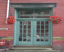 This image provides a view of the paired wood doors with glass panels below the two paned, rectangular transom window and the sandstone lintel, 2005.; City of Saint John