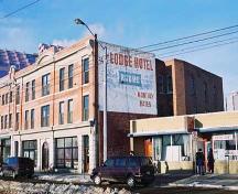 This image illustrates the front, south-facing, primary brick-clad facade of the building showing the storefronts at ground level with two floors of residential uses above. Also illustrated is a large painted sign on the east facade.; City of Edmonton, 2004