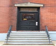 This image provides a view of the entry consisting of a pedimented, sandstone lintel above a stained glass transom window and a wood door with a glass panel, 2005; City of Saint John
