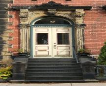 This image provides a view of the entry consisting of an elaborate sandstone entablature supported by brackets, fluted pilasters with Corinthian capitals, Roman arch transom window and paired wood doors with glass panels, 2005; City of Saint John