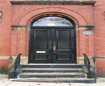 This image provides a view of the Roman arched entrance with brick pilasters crowned by Doric sandstone capitals and paired wood doors, 2005
; City of Saint John