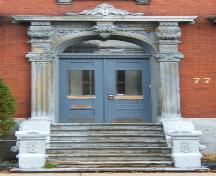 This image provides a view of the entry with the elaborate sandstone entablature supported by brackets and fluted pilasters with Corinthian capitals above a Roman arched transom window and paired wood doors with glass panels, 2005; City of Saint John