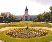 Vue générale de l'édifice de l'Assemblée législative, qui montre sa pelouse, ses massifs floraux, ses arbustes et ses arbres entourés et traversés d'allées de promenade et de routes, 1998.; Parks Canada Agency / Agence Parcs Canada, 1998.