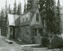 Side view of Building 3, showing the gabled roof, with one stone chimney and the cedar-shingles laid in staggered lines to give a random roof pattern, 1985.; Parks Canada Agency / Agence Parcs Canada, 1985.