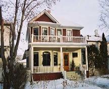 This image illustrates the two storey south and west wood-clad facades with hipped-roof and front gable over two-storey bay window, central chimney, full-width main floor veranda and open balcony above and side bay window with hipped-roof. (March 2004); City of Edmonton, 2004