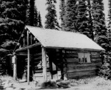 Corner view of the Warden's Cabin at Eva Lake, showing its walls, raised in horizontal logs, dove-tailed at the corners and pierced by a plank door and two multi-paned fixed sash windows, 1986.; Parks Canada Agency / Agence Parcs Canada, 1986.
