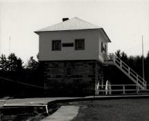 Vue du blockhaus, qui montre la volumétrie carrée, sur deux étages, coiffée d’un toit pyramidal, 1989.; Agence Parcs Canada / Parks Canada Agency, 1989.