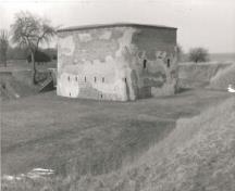 Vue de la Tour, qui montre son emplacement sur un terrain plat gazonné à l’intérieur du remblai de terre du fort, 1989.; Agence Parcs Canada / Parks Canada Agency, 1989.