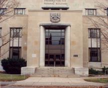 View of the front of the Government of Canada Building, showing its monumental entrance, which consists of, a simplified version of a giant order in marble, resembling engaged columns, 1991.; Department of Public Works / Ministère des Travaux publics, 1991.