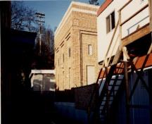 Vue du poste de transformateurs, qui montre sa construction en brique, son toit plat, et sa fondation en béton au contour bien délimité, 1991.; Travaux publics Canada / Public Works Canada, 1991.