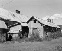 Vue en angle de l’atelier du ferblantier, qui montre la forme rectangulaire simple, le toit à deux versants, le parement et la toiture en tôle ondulée et le ventilateur de faîte, 1988.; Agence Parcs Canada / Parks Canada Agency, 1988.