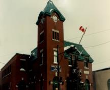 Façade du manège militaire, qui montre le métal peint utilisé pour la toiture à nervures et la tour d’horloge de quatre étages de hauteur, 1989.; Department of National Defence / Ministère de la Défense nationale, 1989.