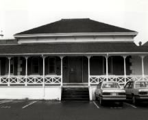 Façade of Building 37, showing its hipped roof, covered verandah, bracketed cornice and the segmentally-arched door and window openings with stone surrounds, 1989.; Ian Doull, Parks Canada Agency / Agence Parcs Canada, 1989.