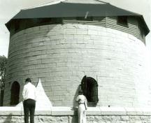 View of the western façade of Murney Martello Tower, showing the limestone walls and the round arched ‘carronade porte’ openings fitted with iron shutters, 1991.; Parks Canada Agency / Agence Parcs Canada, 1991.