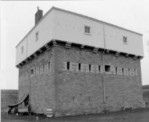 Corner view of the Blockhouse, showing the centrally placed gun ports of the second floor, each flanked by four single-rifle loopholes trimmed with lighter coloured stonework, 1991.; Parks Canada Agency / Agence Parcs Canada, 1991.