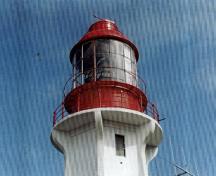 Vue en détail du phare, où l'on peut apercevoir la lanterne cylindrique en métal, surmontée d’un aérateur en dôme et d’une girouette, 1994.; Canadian Coast Guard / Garde côtière canadienne, 1994.