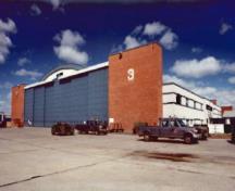 Vue générale du Hangar 3 démontrant les tours massives rectangulaires dans lesquelles se logent les portes d'acier coulissantes, les ailes à bureaux de deux étages à toit plat situées le long des deux côtés du hangar, 1999.; Department of National Defence / Ministère de la Défense nationale, 1999.