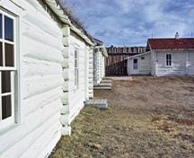Vue de côté de la maison du commissaire (droite) avec autres édifices similaires (gauche) qui démontrent la fondation en béton et les murs extérieurs peints en blanc qui sont faits de rondins écorcés de diamètre semblable posés à l’horizontale, 1972.; Parks Canada Agency / Agence Parcs Canada, 1972.