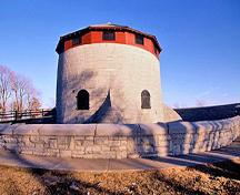 General view of Murney Tower showing the squat round massing of the tower with hive-like caponiers at the base and a conical roof as well as its symmetry, with the circumference and height of the tower in equal proportion, 1994.; Parks Canada Agency/ Agence Parcs Canada, J. Butterill, 1994.