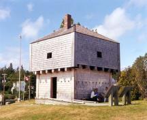 Corner view of St. Andrews Blockhouse, showing the squat square profile, pyramidal roof, entrance and the artillery gunports, machicolation loopholes and musketry holes, 1998.; Parks Canada Agency/Agence Parcs Canada, 1998.