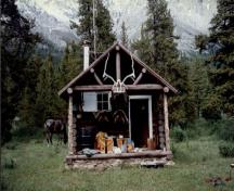 Front elevation of the Topaz Warden Patrol Cabin, showing the gable roof and log-framed open porch with trussed-purlin supports and posts at the gable end, 1997.; Parks Canada Agency / Agence Parcs Canada, 1997.