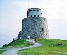 General view of the Carleton Martello Tower showing the exterior walls built of rubble masonry which inclines slightly inwards as it rises to a parapet wall which is in turn topped by a two-storey concrete observation post, 1983.; Parks Canada Agency / Agence Parcs Canada, J. Steeves, 1983.