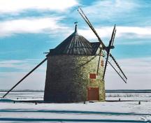 Side view of Round Stone Windmill and House, showing the tall cylindrical three-and-a-half storey massing of the mill under a high conical cap pierced by triangular dormers, 1995.; Parks Canada Agency / Agence Parcs Canada, 1995.