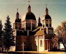 Vue générale de l'église catholique ukrainienne de la Résurrection, qui montre ses tours compactes avec des toits à multiples niveaux et quatre dômes secondaires disposés autour d'un dôme central, 1996.; Parks Canada Agency / Agence Parcs Canada, 1996.