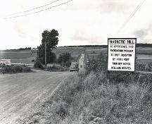 Le signe dans cette photo de vers 1940 dit, «MAGNETIC HILL:  TO APPRECIATE THIS PHENOMENON PROCEED TO SPOT INDICATED BY WHITE POST TURN OFF MOTOR RELEASE BRAKES»; Moncton Museum