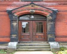 Cette image montre l'entrée comprenant un fronton, des pilastres cannelés avec chapitaux corinthiens, un imposte à arche segmentée et des portes doubles en bois avec panneaux en verre, 2005; City of Saint John