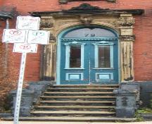 This image provides a view of the entry consisting of an elaborate sandstone entablature supported by brackets, fluted pilasters with Corinthian capitals, a Roman arch transom window and paired wooden doors with glass panels, 2005; City of Saint John