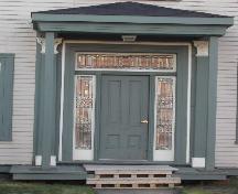 Cette image montre l'entrée centrale avec un toit en croupe, pilastres corinthiens, une imposte en vitrail, des fenêtres latérales et une porte en bois à panneaux de bois, 2006; City of Saint John