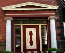 This image shows the entrance with  a pedimented entablature, ionic columns, a wooden door, a transom and sidelights; City of Saint John