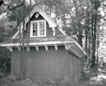 View of the façade of the Wood Shed, showing the gable roof with overhanging and flared eaves supported by large brackets, 1984.; Parks Canada Agency / Agence Parcs Canada, Robert Hunter, 1984.