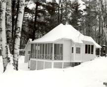 Vue général du chalet des invités, qui montre la véranda fermée et la volumétrie de plain-pied coiffée d’un toit en croupe percé par une cheminée.; Parks Canada Agency / Agence Parcs Canada.