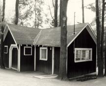 View of the façade of the Tool Shed, showing the one-storey massing of the L-shaped structure with a shingled gable roof with exposed rafter ends, 1984.; Parks Canada Agency / Agence Parcs Canada, M. Trépanier, 1984.