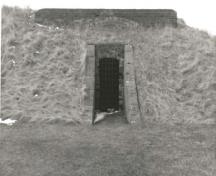 View of the west Powder Magazine, showing the protected entrance flanked by brick sidewalls that slope down to ground level, 1989.; Parks Canada Agency / Agence Parcs Canada, 1989.