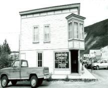 View of the main entrance to Harrington's Store (15), showing the square-sided oriel window placed at the salient angle, the richly ornamented double front door, and the painted cove siding, 1987.; Department of the Environment / Ministère de l'Environnement, 1987.