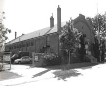 Corner view of the Armoury in Brampton, showing its load-bearing exterior walls clad in red brick with engaged pilasters and detailed with flat-voussoired windows, 1990.; Department of National Defence / Ministère de la Défense nationale, 1990.