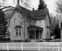 Vue de la façade sud de la maison Bethune, qui montre le bardage à clins des murs extérieurs, l’arrangement symétrique des fenêtres, les baies vitrées et la véranda, 1989.; Agence Parcs Canada / Parks Canada Agency, 1989.