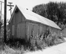 Corner view of the Garage and Fire Hall, showing its rectangular shape, its gable roof, its corrugated metal siding and roof covering, and its wood-frame structure, 1988.; Parks Canada Agency / Agence Parcs Canada, 1988.