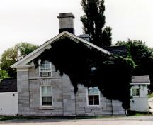 Corner view of RMC Building 2, showing the picturesque qualities of its design and form which complement a domestic garden, picket fence and mature trees, 1993.; Parks Canada Agency / Agence Parcs Canada, 1993.