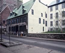 Corner view of the Maillou House showing its exterior masonry walls, the dressed stone of the door and widow frames and string course between the two storeys, and the clapboard cladding of the northeast gable, 1985.; Parks Canada Agency / Agence Parcs Canada, A. Guindon, 1985.