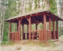 General view of the Pyramid Lake Island Picnic Shelter showing the south elevation and the principles of rustic architecture such as the peeled log frame construction, the vertical log half-height walls, and the wood shingle roof, 1997.; Parks Canada Agency / Agence Parcs Canada, 1997.