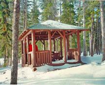 General view of the Pyramid Lake Island Picnic Shelter's east elevation demonstrating the compatibility of the building's rustic form, natural materials and rustic detailing with the picturesque wilderness setting, 1997.; Parks Canada Agency / Agence Parcs Canada, 1997.