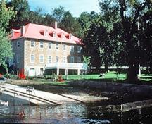 Vue en angle du côté riverain de la caserne de Carillon montrant son emplacement au bord de la rivière des Outaouais, près du canal de Carillon, 1989.; Parks Canada Agency/ Agence Parcs Canada, 1989.