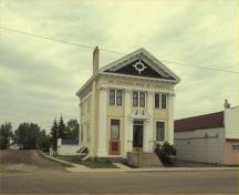 General view of the Canadian Bank of Commerce, showing its neo-classical style, evident in its three-bay facade with classical decorative treatment, particularly the pedimented front gable with bulls-eye window and heavy dentilled cornice, fluted pilaster; Parks Canada Agency / Agence Parcs Canada, 1990.