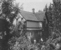 Photo prise de l'extérieur du bâtiment D1, qui montre le volume asymétrique de ce bâtiment de deux étages à plan en L, coiffé d’un toit à deux versants à pente raide et percé d’une cheminée, 1971.; CIHB/IBHC, Parks Canada/Parcs Canada, 1971