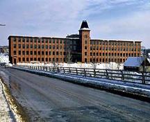 General view of Marysville Cotton Mill, showing its four-storey main elevation with central tower and the imposing scale of the mill building in relation to the surrounding buildings, 1995.; Parks Canada Agency / Agence Parcs Canada, J. Butterill, 1995.