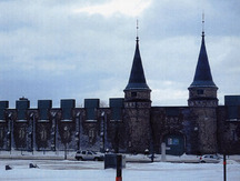 La façade symétrique avec porte centrale voûtée flanquée de tours circulaires à pinacle devant le champ de parade.; Parks Canada | Parcs Canada